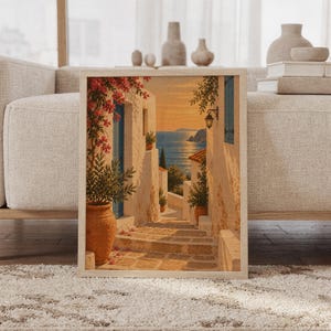Mediterranean coastal village wall art print featuring warm golden sunset tones, ivory stone buildings, weathered blue doors, terracotta pots, and cascading pink bougainvillea framing a shimmering sea view, framed and leaning against a beige sofa on a textured rug in a bright Scandinavian living room.