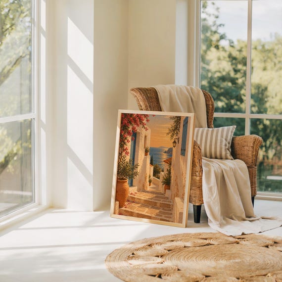 Mediterranean coastal village wall art print leaning on the floor beside a rattan armchair and jute rug in a bright sunlit room.