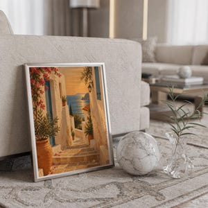 Mediterranean coastal village wall art print in a silver frame leaning against a sofa beside a decorative marble sphere.