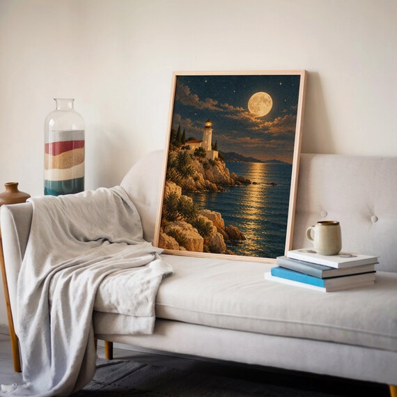 Moonlit lighthouse wall art print leaning on a cream sofa against a beige wall beside books and a mug.
