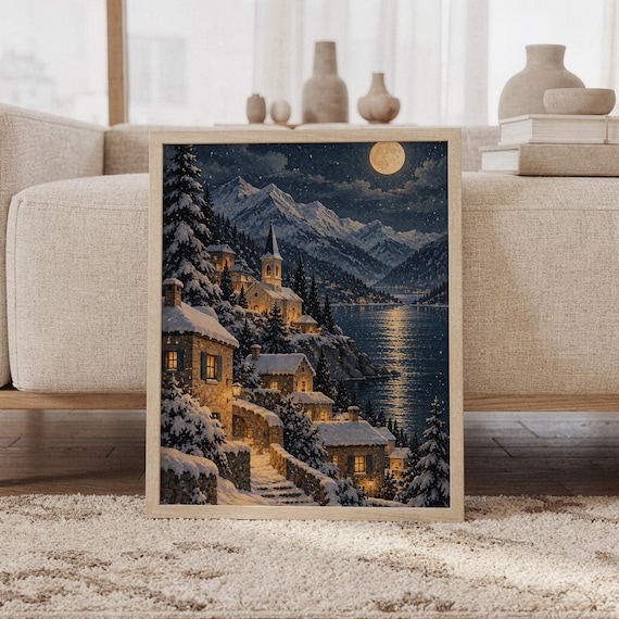 Snowy mountain village wall art print featuring deep navy skies, icy blue mountains, snow-covered stone cottages, glowing amber windows, and a golden full moon in a vintage storybook landscape style, framed and leaning against a beige sofa on a textured rug in a bright Scandinavian living room.