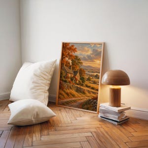 Autumn cottage landscape wall art print leaning on a herringbone wood floor beside cream cushions and a wooden mushroom lamp.