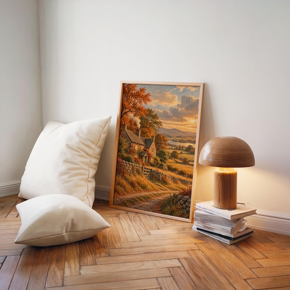 Autumn cottage landscape wall art print leaning on a herringbone wood floor beside cream cushions and a wooden mushroom lamp.