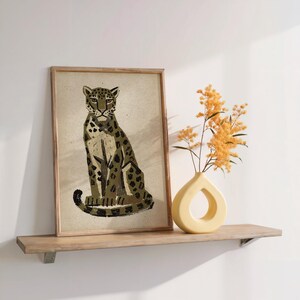 A framed print of a sitting leopard with dark green and brown spots on a light background, on a light wood shelf next to a yellow vase with dried yellow flowers.