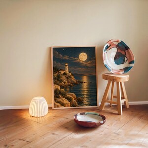 Moonlit lighthouse wall art print leaning against a warm beige wall on a wooden floor beside a stool and a glowing lamp.
