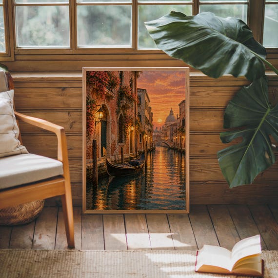 Venice canal sunset wall art print leaning against warm wood paneling beside a wooden armchair and a monstera plant.