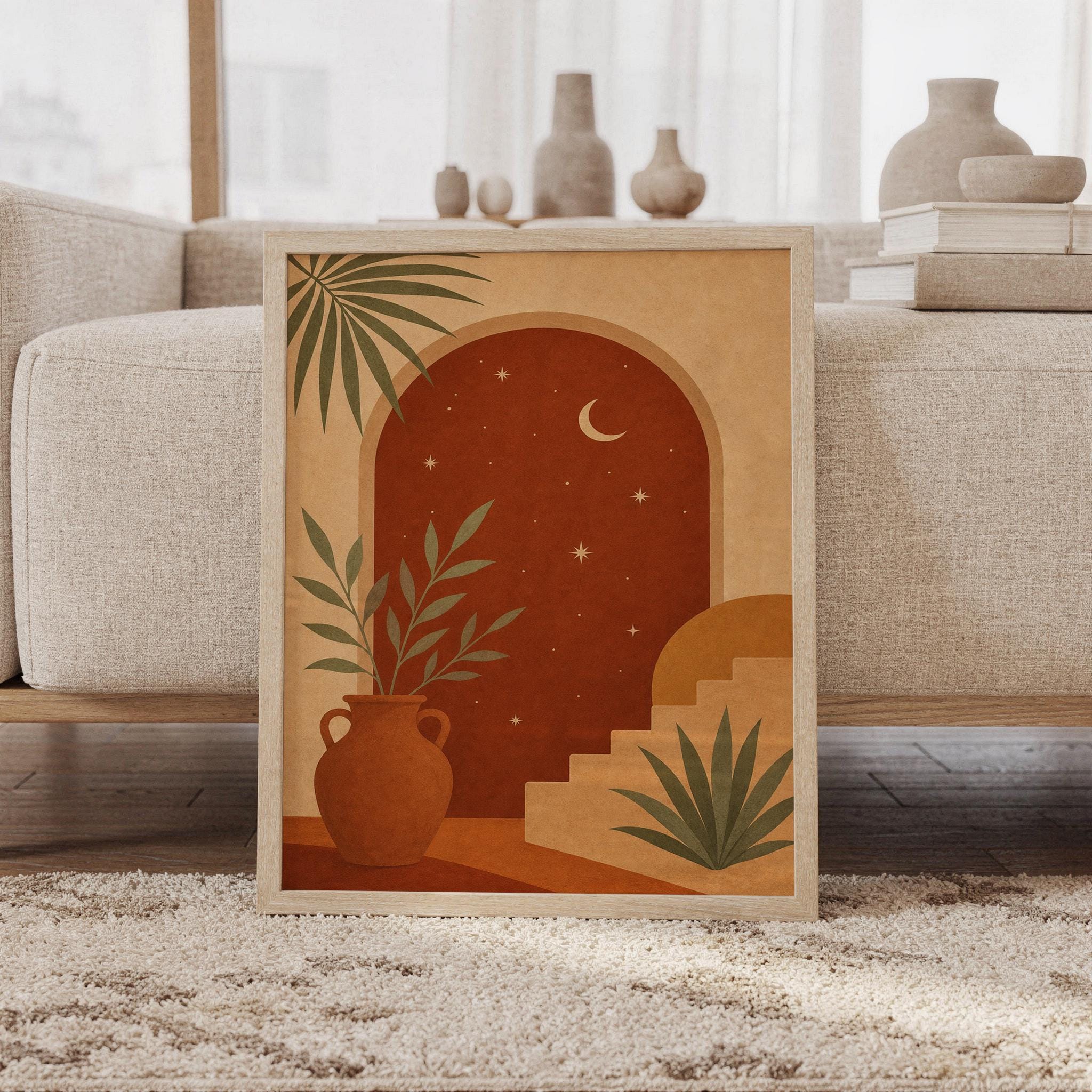 Celestial arch wall art print of a terracotta arched doorway under a night sky with a crescent moon and small stars, curved stairs, a clay vase and palm leaves, framed and leaning against a beige sofa on a textured rug in a bright Scandinavian living room.