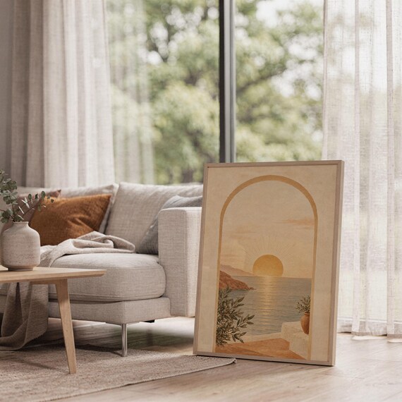 Mediterranean sunset arch print leaning on the floor beside a cream armchair next to large garden windows.