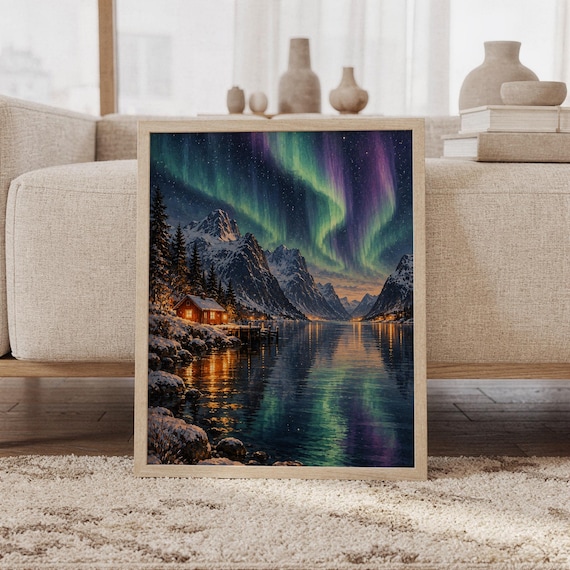 Aurora borealis mountain landscape wall art print featuring luminous green, teal, violet, and blue northern lights above snow-covered mountains, a glowing cabin, and a reflective alpine lake, framed and leaning against a beige sofa on a textured rug in a bright Scandinavian living room.