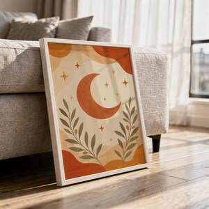 Terracotta crescent moon and olive branch print in a white frame leaning against a gray sofa on a sunlit wooden floor.