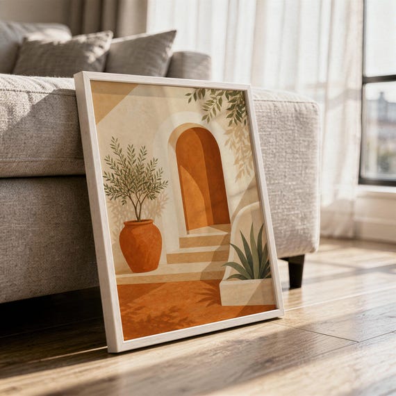 Mediterranean terracotta stairway arch print in a white frame leaning against a gray sofa on a sunlit wooden floor.