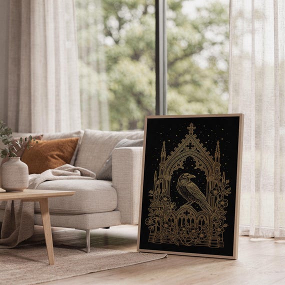 Gothic raven wall art print leaning on the floor beside a cream armchair next to large garden windows.