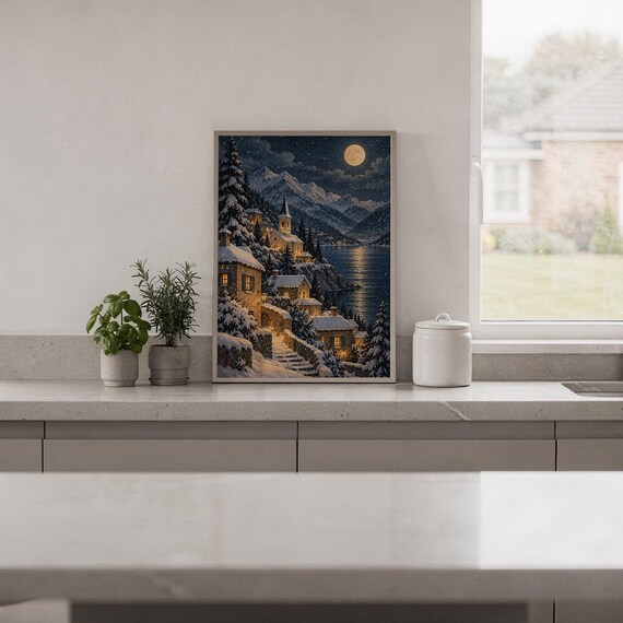 Snowy mountain village wall art print standing on a white kitchen counter beside potted herbs.