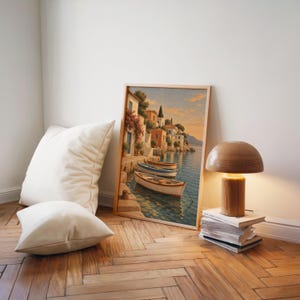 Mediterranean coastal village wall art print leaning on a herringbone wood floor beside cream cushions and a wooden mushroom lamp.