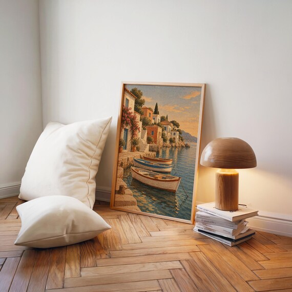 Mediterranean coastal village wall art print leaning on a herringbone wood floor beside cream cushions and a wooden mushroom lamp.