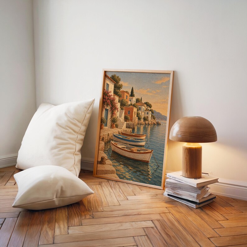 Mediterranean coastal village wall art print leaning on a herringbone wood floor beside cream cushions and a wooden mushroom lamp.
