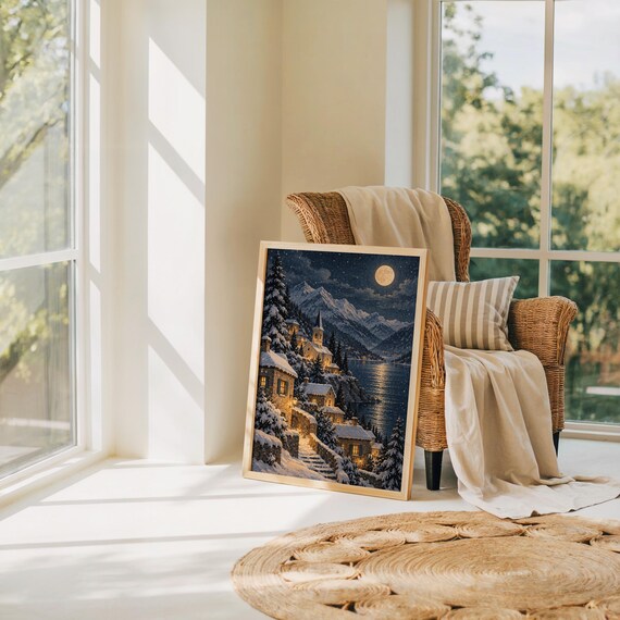 Snowy mountain village wall art print leaning on the floor beside a rattan armchair and jute rug in a bright sunlit room.