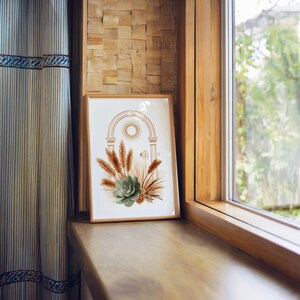 Boho arch botanical art print framed in natural light wood resting on a wide wooden window sill in a sunlit room, overlooking outdoor greenery with soft sunlight filtering through sheer curtains for a serene aesthetic environment.
