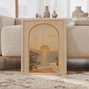 Mediterranean sunset arch wall art print of a glowing sun setting over a calm sea seen through an arch, with coastal cliffs and potted branches in sand, ochre, terracotta and olive tones, framed and leaning against a beige sofa on a textured rug in a bright Scandinavian living room.