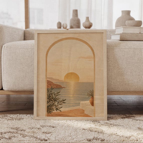 Mediterranean sunset arch wall art print of a glowing sun setting over a calm sea seen through an arch, with coastal cliffs and potted branches in sand, ochre, terracotta and olive tones, framed and leaning against a beige sofa on a textured rug in a bright Scandinavian living room.
