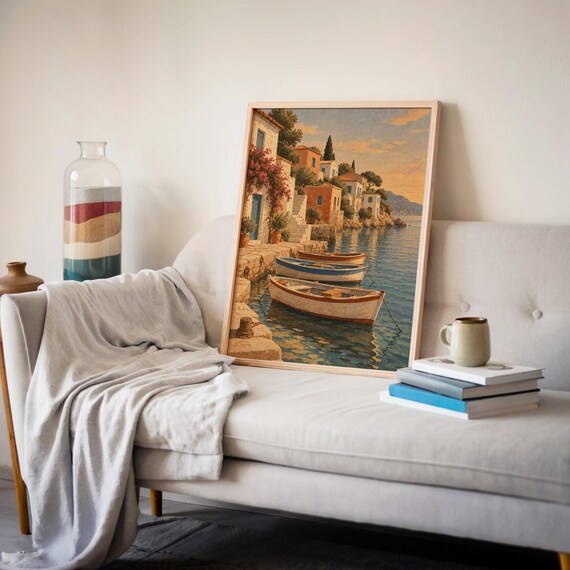Mediterranean coastal village wall art print leaning on a cream sofa against a beige wall beside books and a mug.