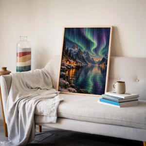 Aurora borealis mountain landscape wall art print leaning on a cream sofa against a beige wall beside books and a mug.