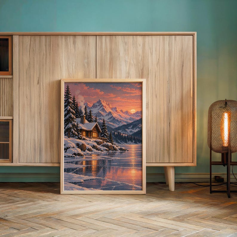 Winter cabin mountain wall art print leaning against a wooden cabinet by a teal wall beside a glowing floor lamp.