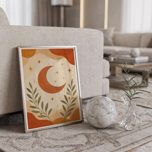 Terracotta crescent moon and olive branch print in a silver frame leaning against a sofa beside a decorative marble sphere.