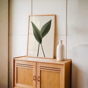A framed print of two dark green leaves on a light background stands on a light brown wooden cabinet, next to a tall white vase.