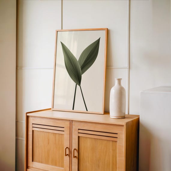 A framed print of two dark green leaves on a light background stands on a light brown wooden cabinet, next to a tall white vase.