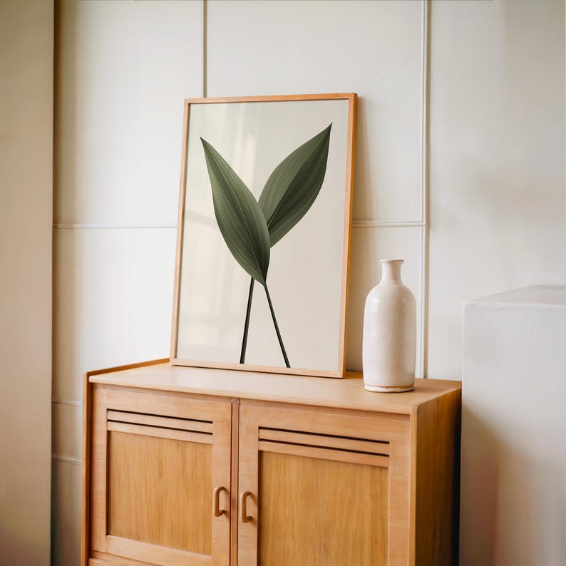 A framed print of two dark green leaves on a light background stands on a light brown wooden cabinet, next to a tall white vase.