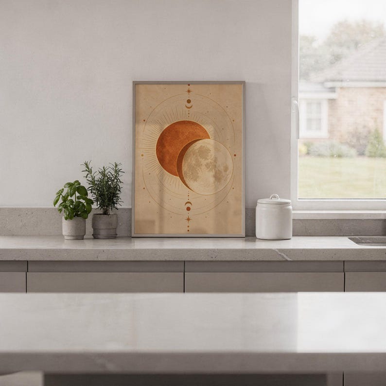 Sun and moon celestial wall art print standing on a white kitchen counter beside potted herbs.