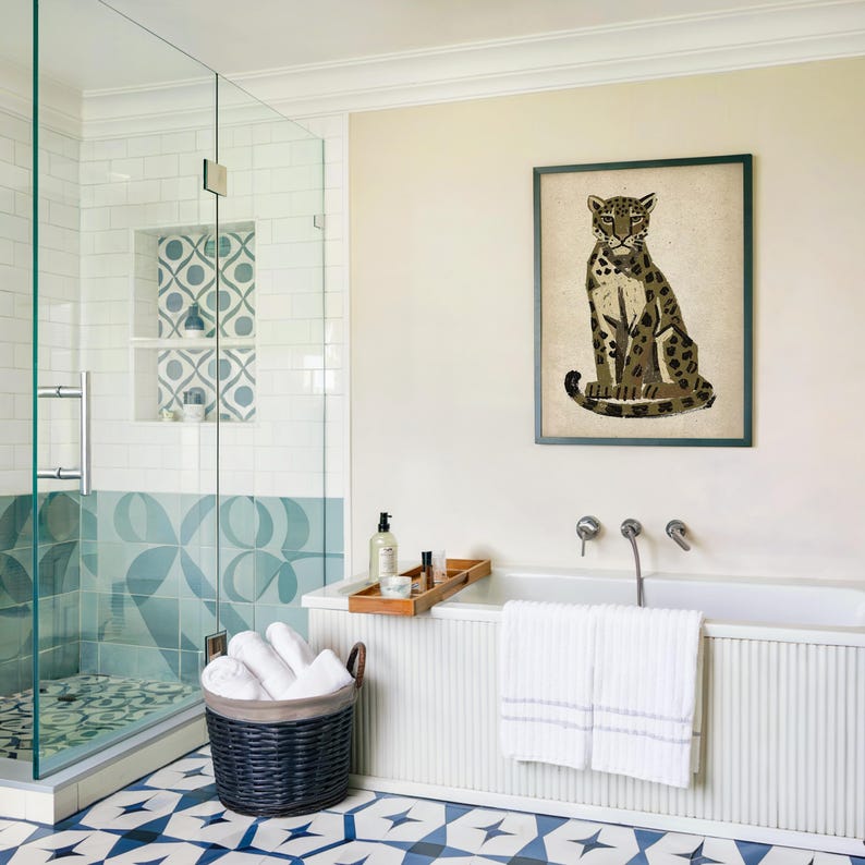 A framed print of a sitting leopard with dark green and brown spots on a light background, hanging on a cream wall above a white bathtub with a wooden tray.