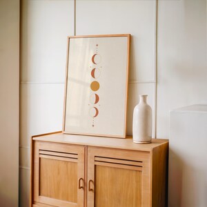 Single terracotta celestial moon phases poster from the set of 3 framed in natural wood resting on top of a vintage wooden sideboard console cabinet beside a tall white decorative vase in a warm room.