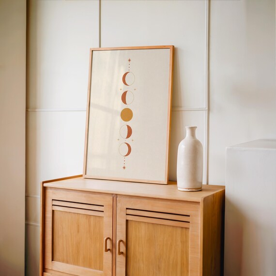 Single terracotta celestial moon phases poster from the set of 3 framed in natural wood resting on top of a vintage wooden sideboard console cabinet beside a tall white decorative vase in a warm room.