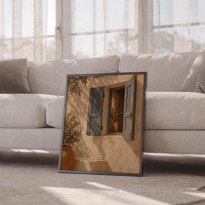 Mediterranean blue shutter wall art print leaning against a white sofa in a bright living room with sheer curtains.