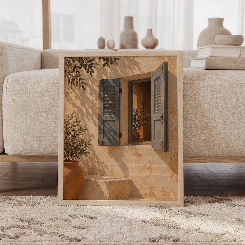 Mediterranean blue shutter wall art print featuring sun-washed cream stone, weathered blue shutters, terracotta pottery, and leafy olive branches in a rustic Mediterranean style, framed and leaning against a beige sofa on a textured rug in a bright Scandinavian living room.
