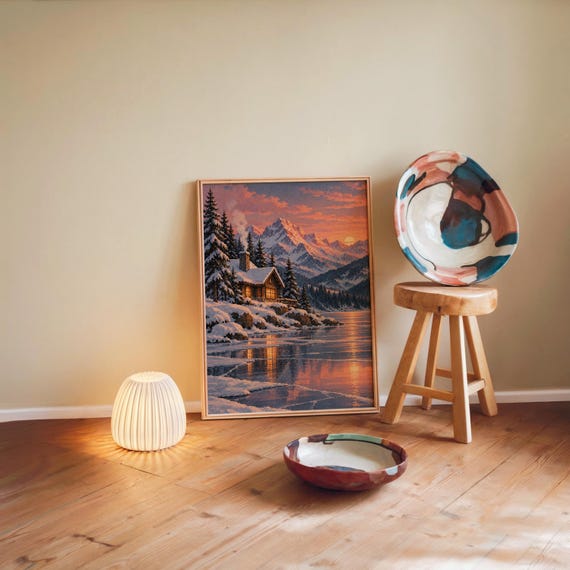 Winter cabin mountain wall art print leaning against a warm beige wall on a wooden floor beside a stool and a glowing lamp.