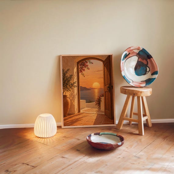 Mediterranean sunset doorway wall art print leaning against a warm beige wall on a wooden floor beside a stool and a glowing lamp.