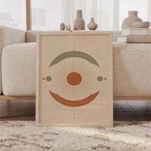 Abstract sun wall art print featuring warm terracotta, muted sage green, and soft cream tones with a radiant sunburst and layered geometric circles, framed and leaning against a beige sofa on a textured rug in a bright Scandinavian living room.