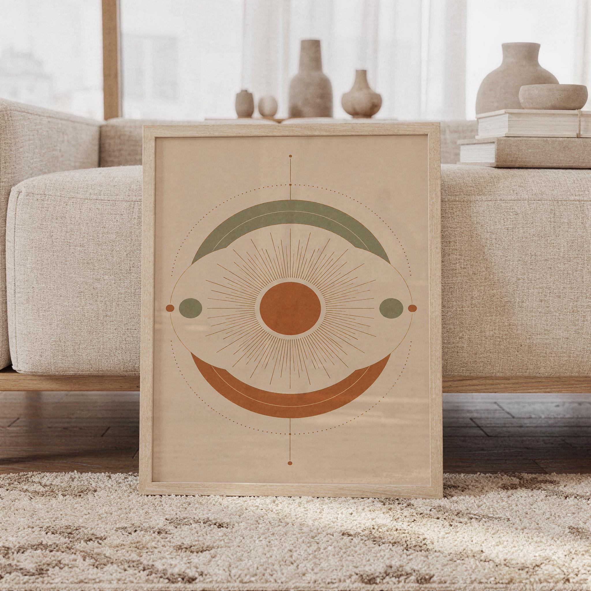 Abstract sun wall art print featuring warm terracotta, muted sage green, and soft cream tones with a radiant sunburst and layered geometric circles, framed and leaning against a beige sofa on a textured rug in a bright Scandinavian living room.