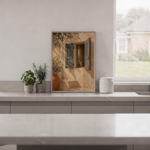 Mediterranean blue shutter wall art print standing on a white kitchen counter beside potted herbs.