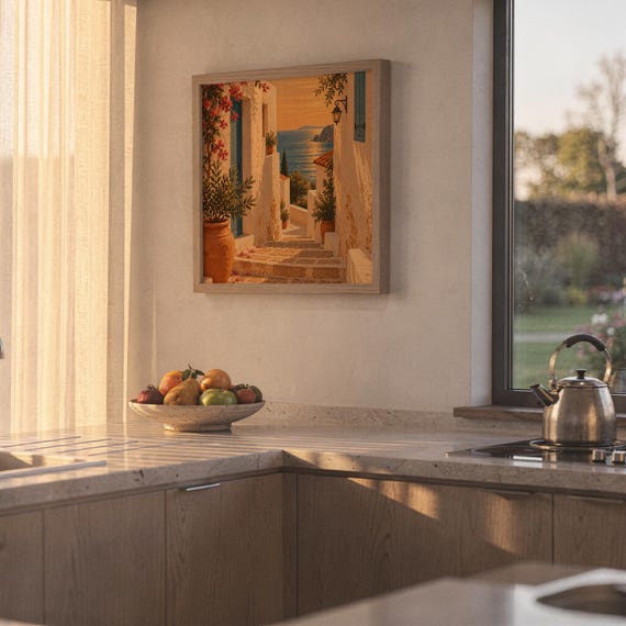 Mediterranean coastal village wall art print hanging above a stone countertop with a fruit bowl in a sunny kitchen.