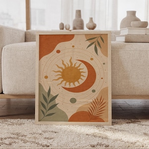 Celestial sun and moon wall art print of a radiant sun and crescent moon with orbiting planets and leaves in terracotta, golden ochre, sage and cream, framed and leaning against a beige sofa on a textured rug in a bright Scandinavian living room.