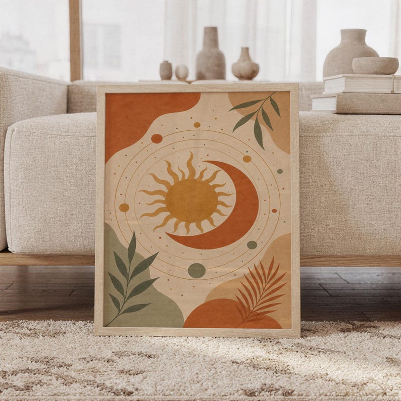 Celestial sun and moon wall art print of a radiant sun and crescent moon with orbiting planets and leaves in terracotta, golden ochre, sage and cream, framed and leaning against a beige sofa on a textured rug in a bright Scandinavian living room.