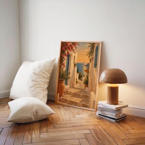 Mediterranean coastal village wall art print leaning on a herringbone wood floor beside cream cushions and a wooden mushroom lamp.