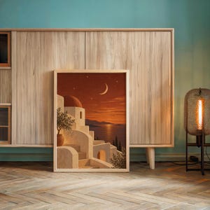 Mediterranean moonlit coastal wall art print leaning against a wooden cabinet by a teal wall beside a glowing floor lamp.