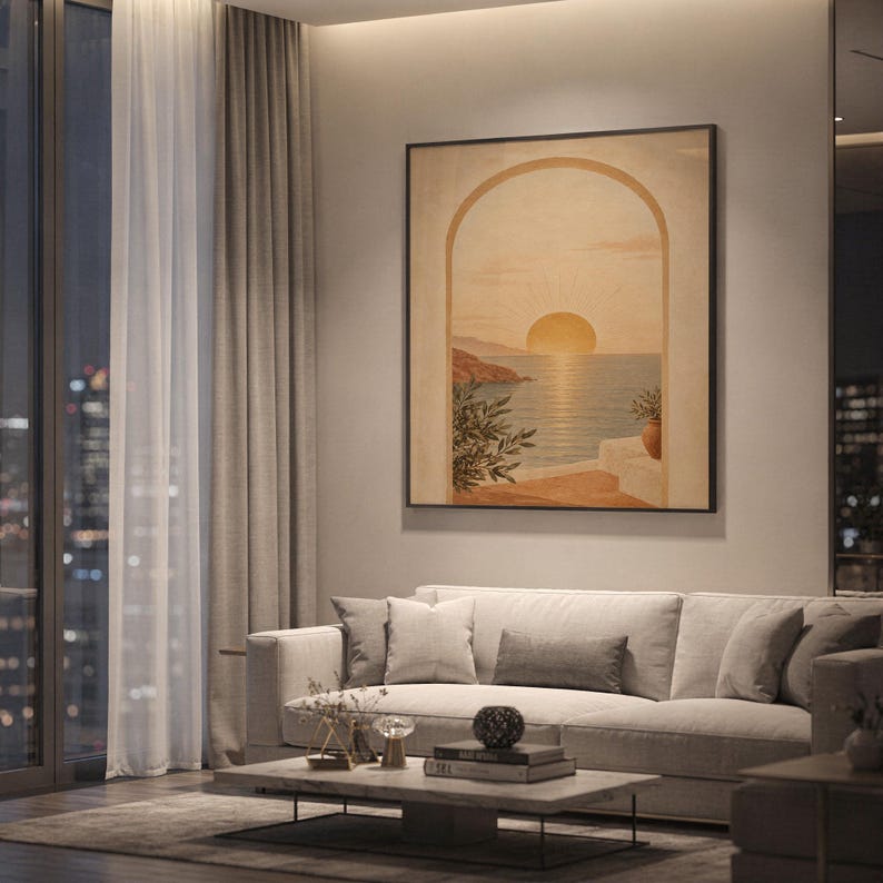 Mediterranean sunset arch print showing a glowing sun setting over a calm sea seen through an arch, with coastal cliffs and potted branches in sand, ochre, terracotta and olive tones, hanging above a light gray sofa in a modern living room with evening city views through tall windows.