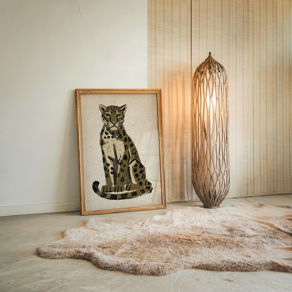 A framed print of a sitting leopard with dark green and brown spots on a light background, leaning against a white wall next to a tall, woven floor lamp and a faux fur rug.