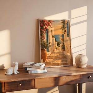 Mediterranean coastal village wall art print leaning on a wooden desk against a warm beige wall in soft window sunlight.